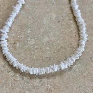 May include: A white shell necklace made of small, flat, white shells. The necklace is strung together with a simple knot.
