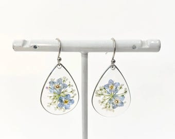Puede incluir: Un par de pendientes en forma de lágrima con ganchos plateados. Cada pendiente presenta una lágrima de resina transparente con un grupo de flores prensadas azules y blancas en su interior.