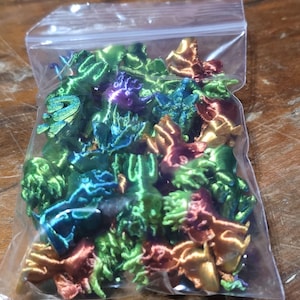 May include: A plastic bag filled with small, colorful, 3D printed objects. The objects are in various shades of green, blue, purple, orange, and gold.