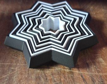 May include: A set of seven black and white star-shaped cookie cutters, nested together on a wooden surface.