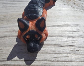May include: A 3D printed figurine of a German Shepherd dog lying down. The figurine is made of orange and black plastic and has a detailed design.