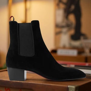 May include: Black suede ankle boot with a pointed toe and a small, block heel. The boot features elastic side panels and a pull tab with red and white accents. The sole has a red accent.