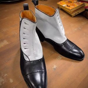 Puede incluir: Botas de cuero bicolor, con puntera y talón negros y parte superior blanca. Las botas tienen un diseño con cordones y una pequeña lengüeta en la parte trasera. El interior es de color naranja cálido. Estilo clásico.