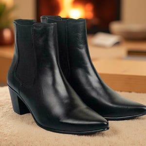 以下が含まれることがあります： 黒い革のアンクルブーツのペア。つま先が尖っており、ブロックヒールが付いています。ブーツには伸縮性のあるサイドパネルが付いています。背景には暖炉と木製の家具があります。ニュートラルな表面に置かれています。