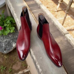 Könnte beinhalten: Ein Paar burgunderfarbene Chelsea-Stiefel aus Leder mit schwarzen elastischen Seiteneinsätzen. Die Stiefel haben eine glänzende Oberfläche und eine spitze Zehenpartie. Die Schuhe werden im Freien auf einer Betonoberfläche präsentiert.