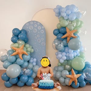 Puede incluir: Una escena de fiesta de cumplea&ntilde;os con globos azules, celeste y verdes, y decoraciones de estrellas de mar naranjas. Un pastel azul con glaseado azul y la palabra "one" frente a un ni&ntilde;o. Un arco azul con dise&ntilde;os de burbujas en el fondo.