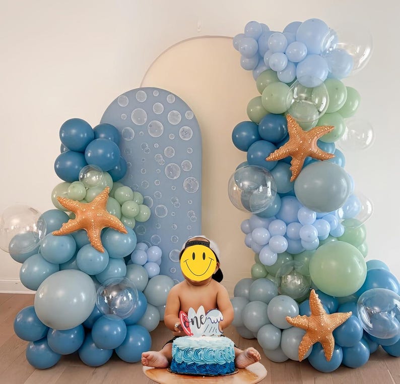 Puede incluir: Una escena de fiesta de cumplea&ntilde;os con globos azules, celeste y verdes, y decoraciones de estrellas de mar naranjas. Un pastel azul con glaseado azul y la palabra "one" frente a un ni&ntilde;o. Un arco azul con dise&ntilde;os de burbujas en el fondo.