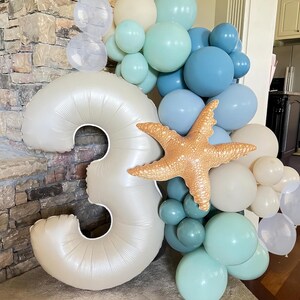 Puede incluir: Un arreglo de globos con un gran n&uacute;mero 3 nacarado, rodeado de globos azules, turquesas y blancos. Un globo de estrella de mar a&ntilde;ade un toque n&aacute;utico a la decoraci&oacute;n de cumplea&ntilde;os.