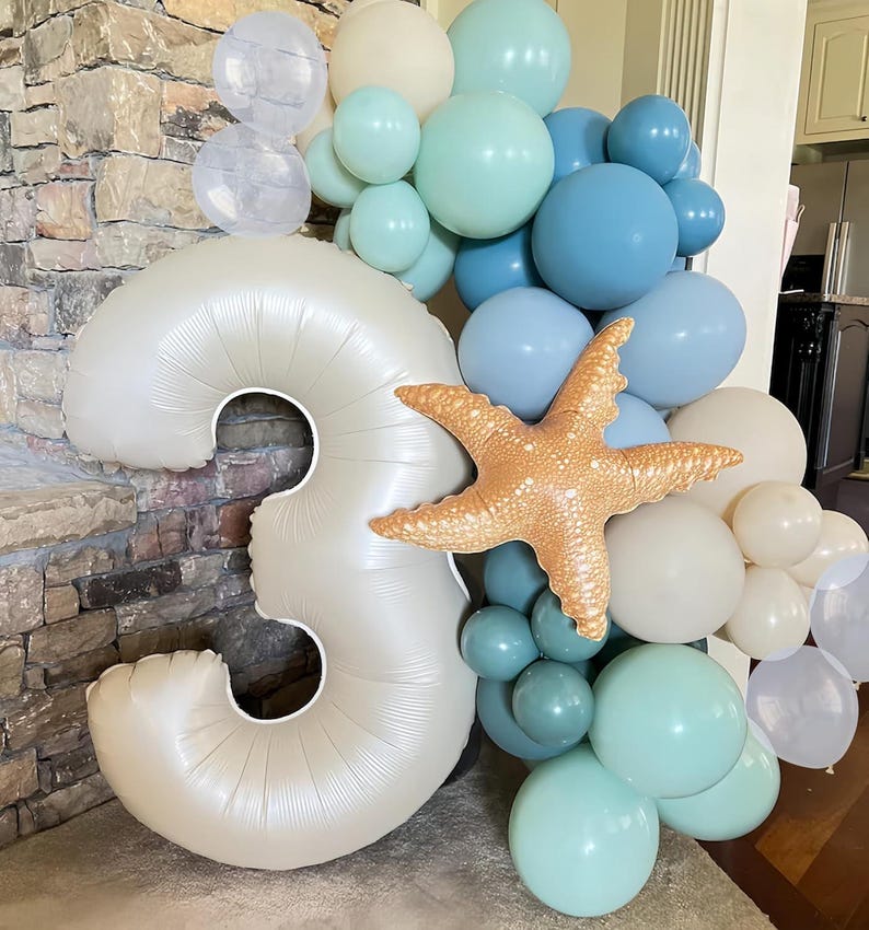 Puede incluir: Un arreglo de globos con un gran n&uacute;mero 3 nacarado, rodeado de globos azules, turquesas y blancos. Un globo de estrella de mar a&ntilde;ade un toque n&aacute;utico a la decoraci&oacute;n de cumplea&ntilde;os.