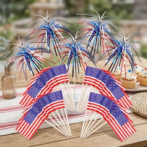 Könnte beinhalten: Eine Sammlung patriotischer Partydekorationen, darunter Miniatur-US-Flaggen auf Holzstäben und sternförmige Aufsätze in Rot, Weiß und Blau. Cupcakes mit Sternenstreuseln sind im Hintergrund zu sehen.