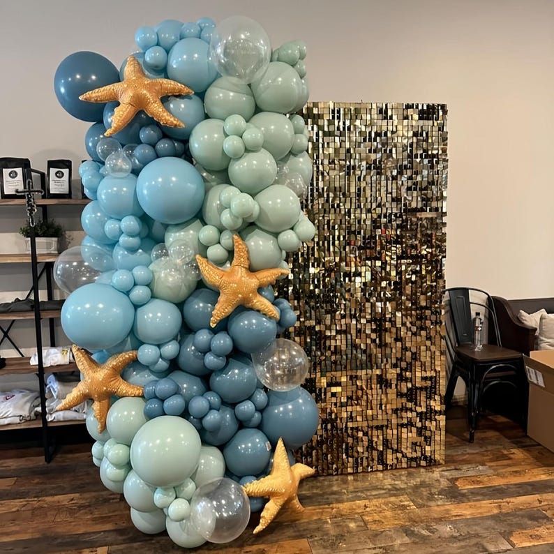 Puede incluir: Un arco de globos en tonos azules y transparentes, decorado con estrellas de mar doradas. El arco est&aacute; junto a una pared de lentejuelas doradas, creando un tel&oacute;n de fondo festivo. Los globos est&aacute;n dispuestos en un dise&ntilde;o en cascada, perfecto para una fiesta.