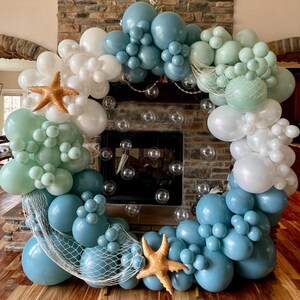 Puede incluir: Una guirnalda de globos circular en tonos azules, blancos y verdes, decorada con estrellas de mar y redes. Los globos est&aacute;n dispuestos en racimos, creando una exhibici&oacute;n festiva y decorativa. Ideal para un evento tem&aacute;tico n&aacute;utico u oce&aacute;nico.