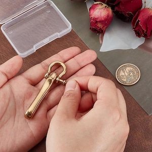May include: A brass-colored, U-shaped clasp with a cylindrical body and a screw closure. The clasp is held in a person's hands, with a clear plastic case in the background. A US one-dollar coin is also visible.