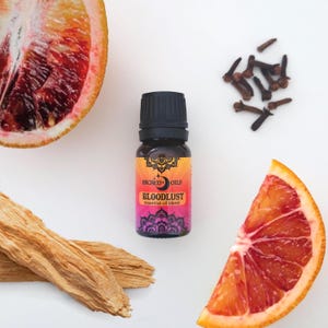 May include: A small brown glass bottle with a black cap, labelled "Sacred Oils Bloodlust essential oil blend". The bottle is surrounded by orange slices, cloves, and a piece of wood.
