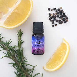 May include: A small brown glass bottle with a black cap, labelled "Sacred Oils Nightfall Essential Oil Blend". The bottle is surrounded by lemon slices, black peppercorns, and rosemary sprigs.