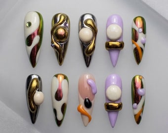 May include: A collection of ten artificial nails with various designs. The nails feature a mix of colors, including gold, silver, white, purple, and orange. Some nails have 3D embellishments, while others have abstract painted designs. The nails are arranged on a white surface.