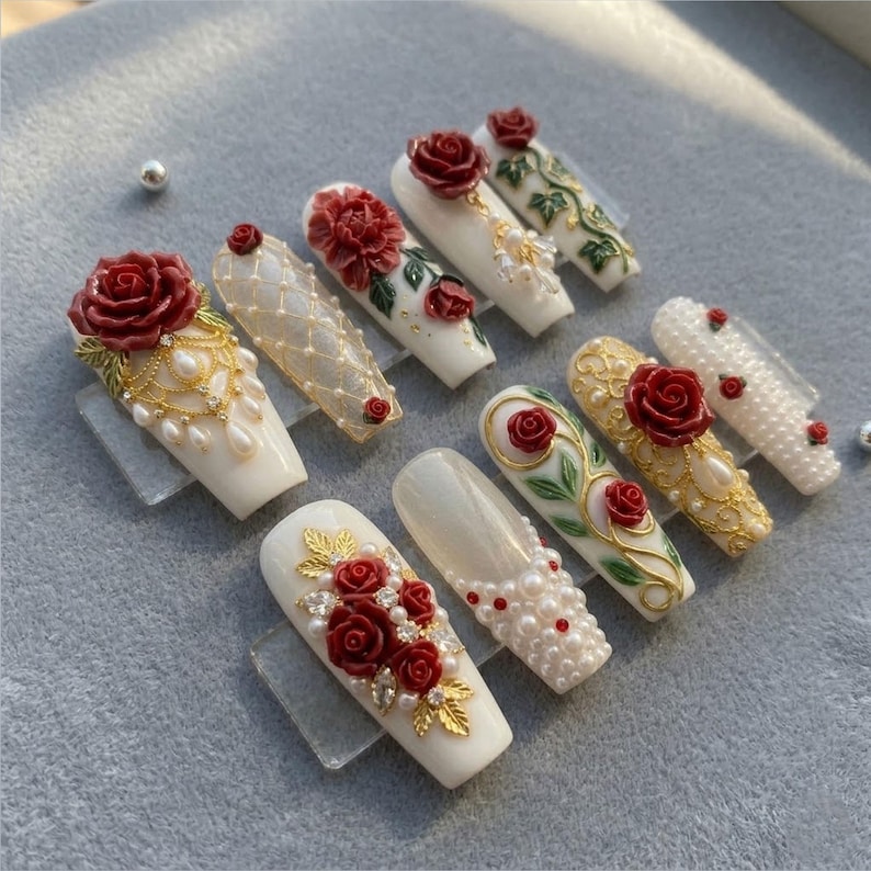 May include: A collection of ten artificial nails, each with a white base and elaborate floral designs. The nails feature red roses, gold accents, pearls, and rhinestones, creating a luxurious and ornate aesthetic.