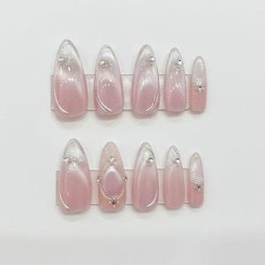 May include: Set of ten artificial nails in a light pink and white ombre design. Each nail features a silver gem accent. The nails are arranged in two rows of five on a clear plastic holder.