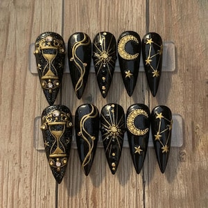 May include: Black press-on nails with gold celestial designs. Each nail features a different design, including an hourglass, sunburst, crescent moon, and constellations. The nails are long and pointed, with a glittery finish.