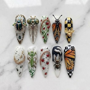 May include: A collection of ten long, pointed fingernails, each uniquely decorated with insect-themed designs. The nails feature detailed artwork of beetles, butterflies, bees, and other insects, with accents of gold, black, and white. The designs include checkered patterns and intricate details.