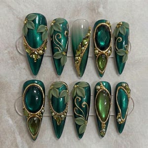 May include: A set of ten stiletto-shaped press-on nails in deep emerald green. The designs feature 3D matte green leaves, gold metallic chains, intricate gold swirls, and faceted dark green and olive-toned resin gemstones with small gold beads.