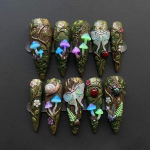 May include: A set of ten long, pointed false nails with a forest theme. Each nail features a unique design with mushrooms, vines, butterflies, ladybugs, and snails in shades of green, brown, and iridescent colors.