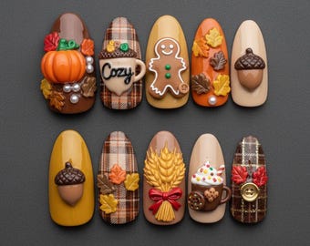 May include: A set of ten press-on nails featuring autumn-themed 3D decorations including a pumpkin, a mug with the text Cozy, a gingerbread person, acorns, plaid patterns, wheat stalks, a hot cocoa mug with whipped cream, and a gold button.