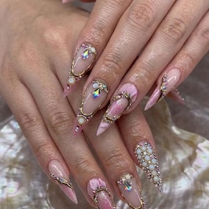 May include: Long, pointed acrylic nails with a light pink base and intricate gold and jewel embellishments. Some nails feature a cluster of small, white pearls, while others have a gold frame with a large, iridescent gem.