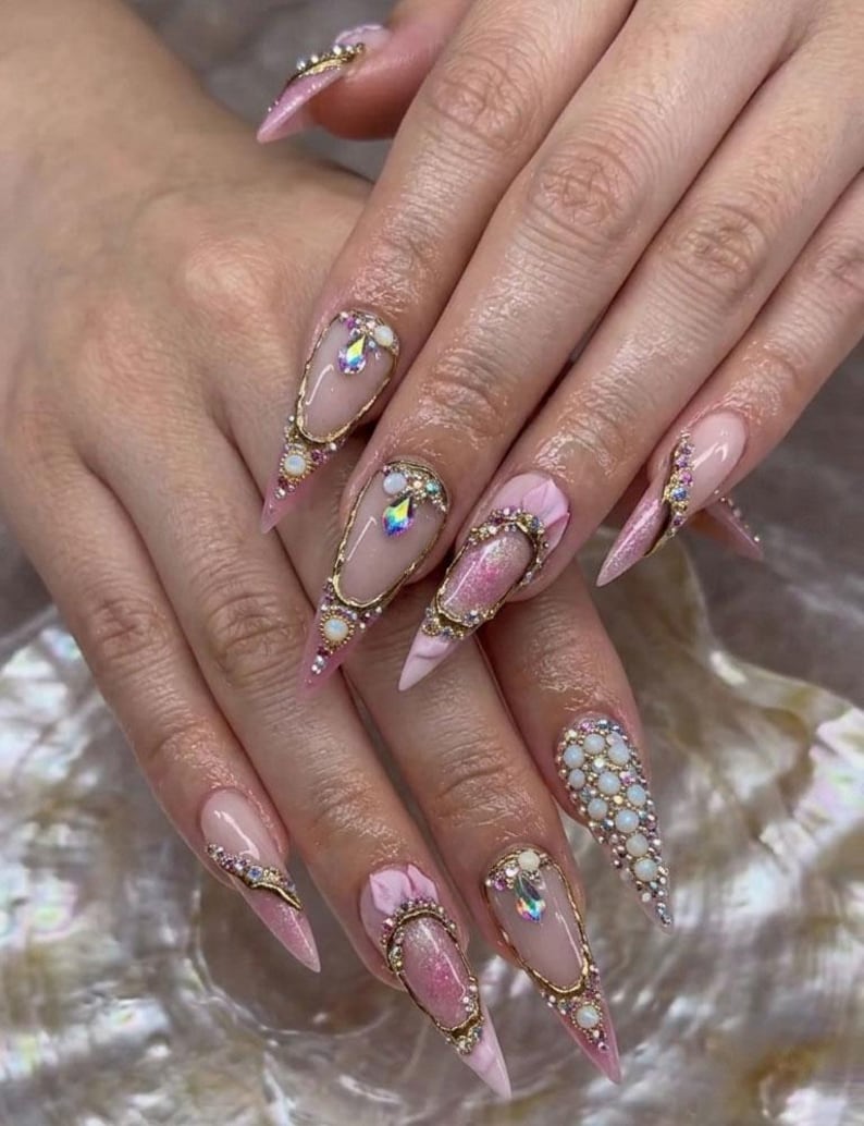 May include: Long, pointed acrylic nails with a light pink base and intricate gold and jewel embellishments. Some nails feature a cluster of small, white pearls, while others have a gold frame with a large, iridescent gem.