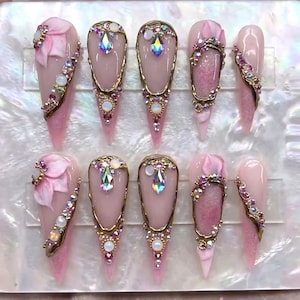 May include: A set of ten long, pointed artificial nails. Each nail is painted a light pink with intricate designs. Some nails feature 3D floral accents, while others have gold-colored frames and sparkling gemstones.