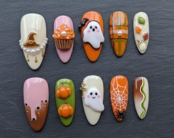 May include: A set of ten almond-shaped press-on nails featuring Halloween designs including a witch hat with pearls, a cupcake, ghosts, orange plaid, autumn leaves, dripping pink and brown polish, pumpkins, a spider web with a spider, and a cross.