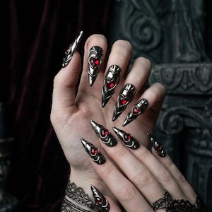 Op de afbeelding: Een set zilverkleurige, puntige nagelhoezen met een hartvormig, rood edelsteenaccent. De nagelhoezen zijn ontworpen om op lange, scherpe klauwen te lijken. De hand is versierd met een zwarte kanten armband en ring.