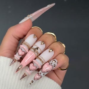 May include: Long stiletto-shaped artificial nails featuring a matte white and glossy pink colour palette. The nails are decorated with metallic gold chains, small gold studs, and raised pink heart-shaped charms. Each nail tapers to a sharp point.