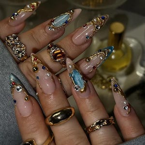 May include: Long, almond-shaped artificial nails with a nude base, adorned with gold accents, pearls, and colorful gemstones. Various gold rings with intricate designs and gemstones are also visible.