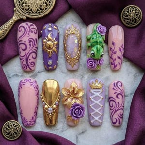 May include: A set of ten press-on nails featuring purple and pink base colours with intricate designs including a 3D green lizard, gold sun, gold crown, floral accents, purple swirls, and a corset-style lace-up pattern with gold and clear rhinestones.