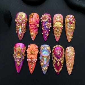 May include: Ten artificial nails, each with a unique, ornate design. The nails are adorned with flowers, seashells, chains, and gems in pink, gold, orange, and purple hues. The nails are displayed against a black backdrop.
