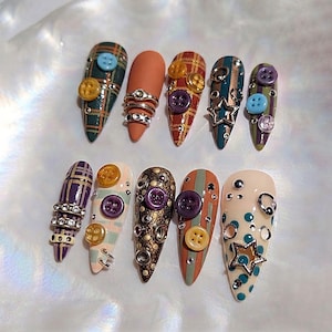May include: A set of ten press-on nails featuring almond-shaped tips decorated with various embellishments including small plastic buttons, metallic star charms, silver rings, and rhinestones. The designs include plaid patterns, solid colors, and polka dots.