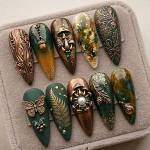 May include: A collection of ten long, almond-shaped artificial nails. Each nail features a unique design, including wood grain, acorns, mushrooms, gold leaf, and botanical patterns. The color palette includes shades of green, brown, and gold, with metallic accents.