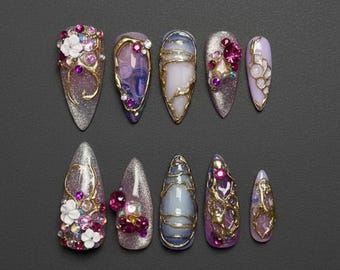 May include: A collection of ten elaborately designed artificial nails. Each nail features a unique design with glitter, gold accents, and various embellishments like flowers and gemstones in shades of pink, purple, and white. The nails are set against a dark gray background.