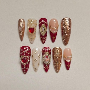 May include: A collection of ten long, almond-shaped artificial nails. The nails feature various designs, including red, gold, and nude colors, adorned with rhinestones, pearls, and intricate gold embellishments. Some nails have heart-shaped gems and ornate frames.