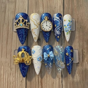 May include: A set of ten long, pointed artificial nails. Designs include a gold crown, a pocket watch, a carriage, a bow, and a glass slipper. The nails feature blue glitter, white lace, and gold accents. Some nails have star and floral details.