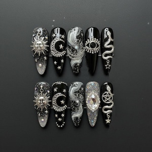 May include: A set of ten long, pointed artificial nails. Each nail features a black base with silver celestial designs, including suns, moons, stars, and snakes. Some nails have a glitter gradient, while others have a large gem or chain accents.
