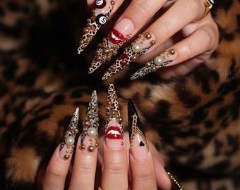 May include: A set of long, pointed acrylic nails with various designs. Some nails feature leopard print, gold embellishments, pearls, and red lips. One nail has a black heart design. The nails are adorned with gold rings.