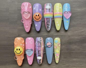 May include: A set of ten long, almond-shaped artificial nails. Each nail features a unique design, including smiley faces, hearts, stars, rainbows, and a planet. The color palette includes pink, orange, purple, yellow, and blue, with clear bubble accents.