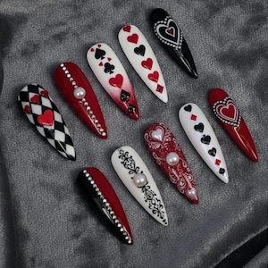 May include: A collection of long, almond-shaped artificial nails with various designs. The nails feature playing card suits, hearts, and decorative elements like rhinestones and pearls. Colors include red, black, white, and silver. The nails are arranged on a gray surface.