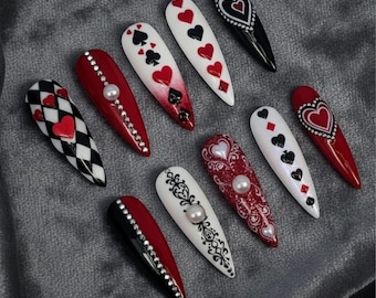 May include: A collection of long, almond-shaped artificial nails with various designs. The nails feature playing card suits, hearts, and decorative elements like rhinestones and pearls. Colors include red, black, white, and silver. The nails are arranged on a gray surface.