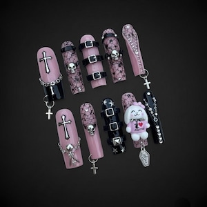 May include: A collection of press-on nails in a gothic style. Designs include crosses, skulls, buckles, and a coffin shape. Colors are black, pink, and silver. Some nails feature chains and charms for added detail.