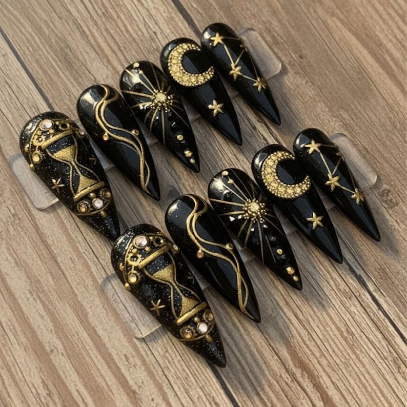 May include: A set of ten black press-on nails with gold celestial designs. The designs include moons, stars, suns, and hourglasses. Each nail is adorned with gold glitter and rhinestones, creating a glamorous and mystical aesthetic.