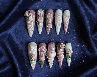 May include: A collection of ten long, pointed artificial nails. Each nail showcases a unique design, including pink roses, bows, and swan figures. The nails are painted in shades of pink, white, and nude, with detailed decorations and embellishments.