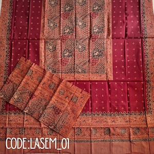 Könnte beinhalten: Ein tiefroter Stoff mit einem Mittelteil aus komplexen schwarzen und roten Blumenmustern, umgeben von einer Bordüre aus kleinen weißen Kreuzmotiven. Das Textil ist gefaltet und zeigt einen Abschnitt mit dichten, dunklen Batikmustern.
